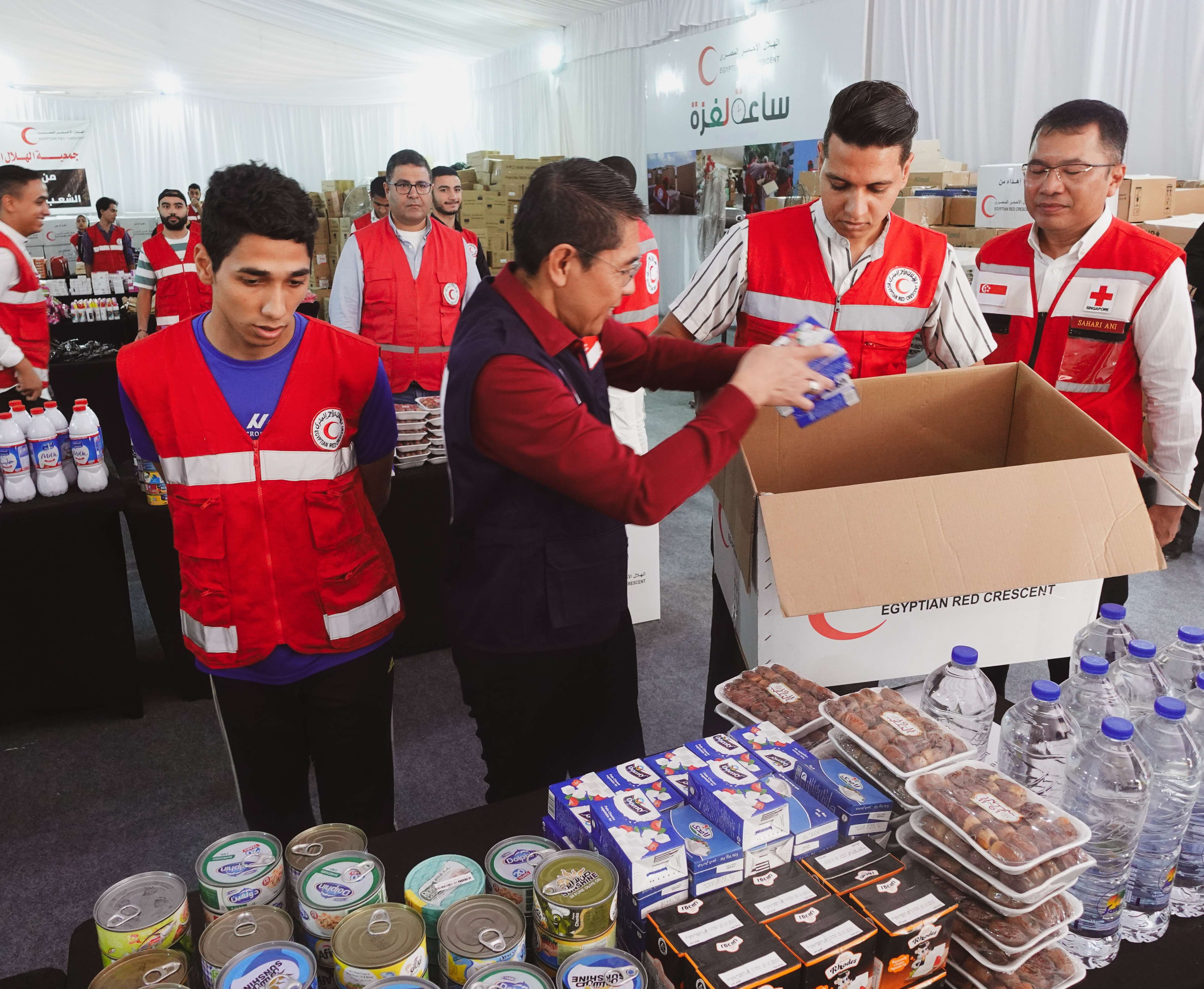 People in red vests packing food items into an "Egyptian Red Crescent" box with water bottles on a table.